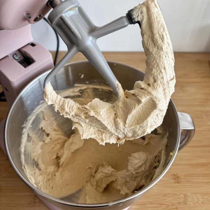 A stand mixer with a paddle attachment holds creamy, light brown batter, with more batter visible in the metal mixing bowl below.