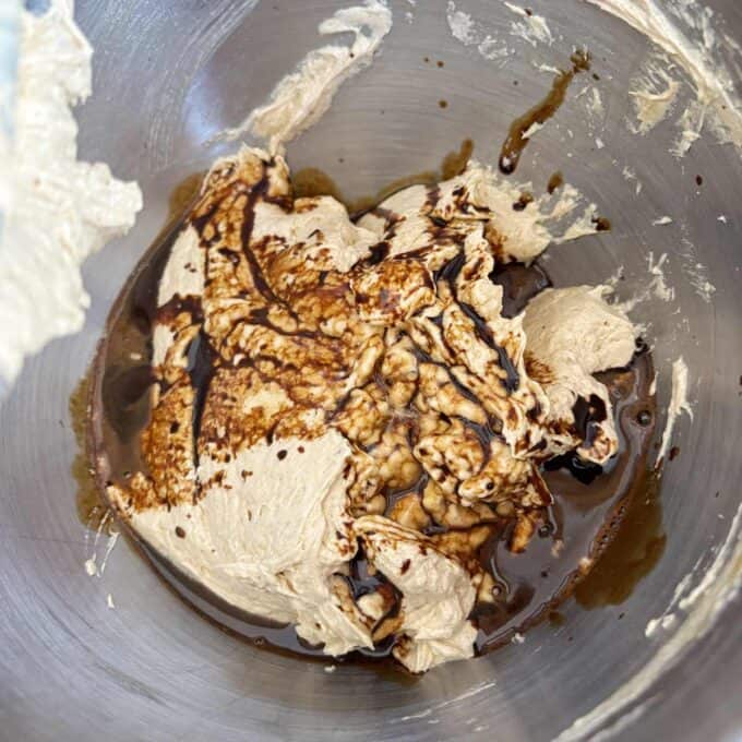A mixing bowl containing light brown whipped mixture with dark liquid, likely vanilla extract, poured over it.