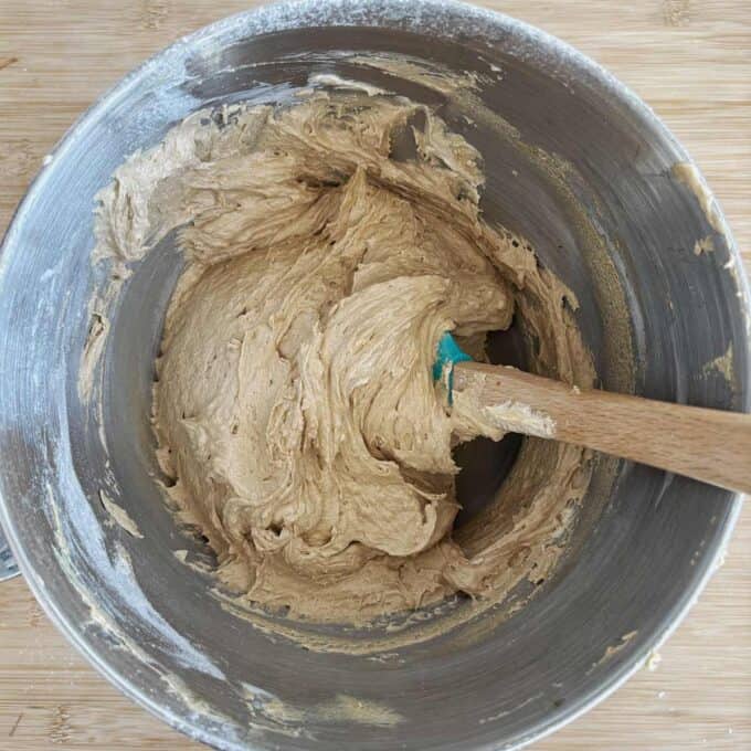 A metal mixing bowl containing thick cookie dough being stirred with a spatula on a wooden surface.