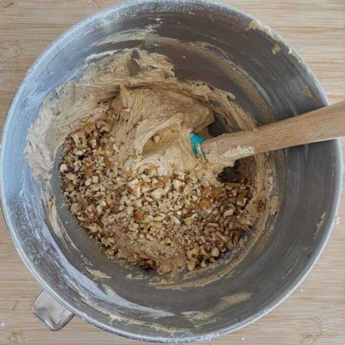 A mixing bowl containing thick brown batter with chopped nuts being stirred in using a wooden spoon.