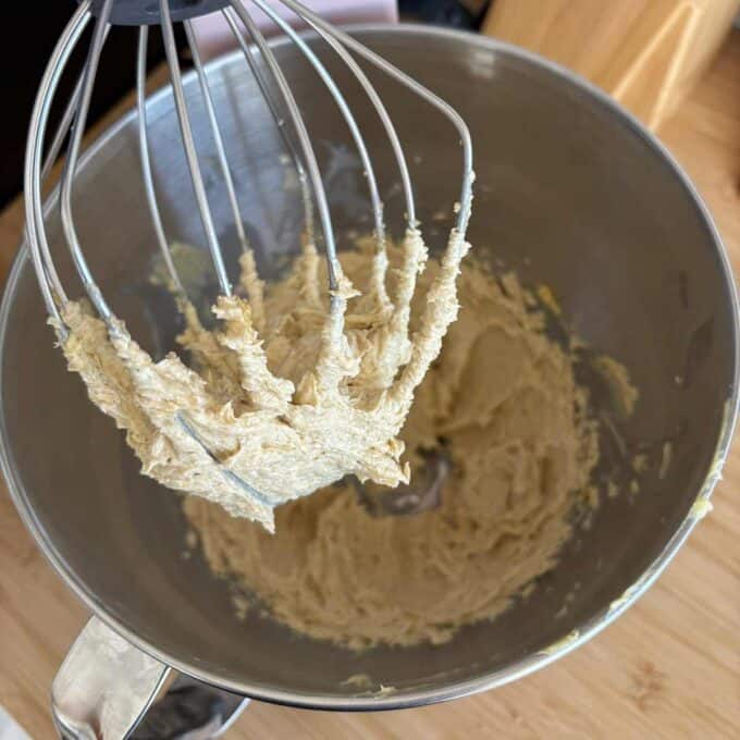 A stand mixer with a whisk attachment coated in thick cookie dough sits above a mixing bowl containing more dough on a wooden surface.