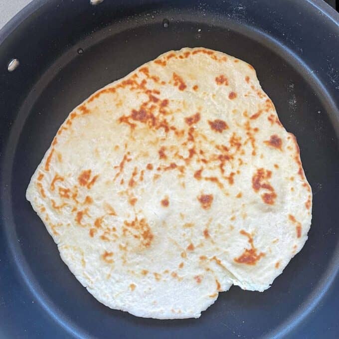 A single flatbread with a golden-brown, uneven surface is cooking in a black nonstick skillet.