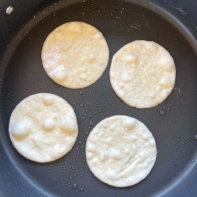 Four small, round pieces of dough are cooking in a frying pan, with visible bubbles forming on their surfaces.