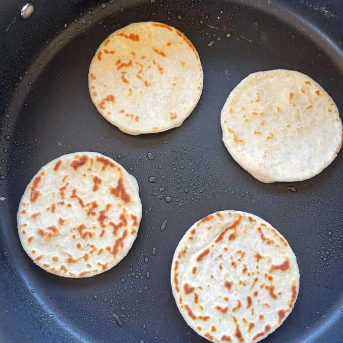 Three small round flatbreads cooking in a frying pan with some droplets of oil visible on the pans surface.