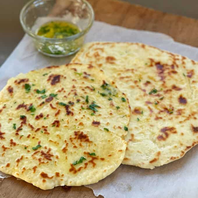 Two pieces of flatbread topped with herbs are placed on parchment paper, with a small glass bowl of herb-infused oil in the background.