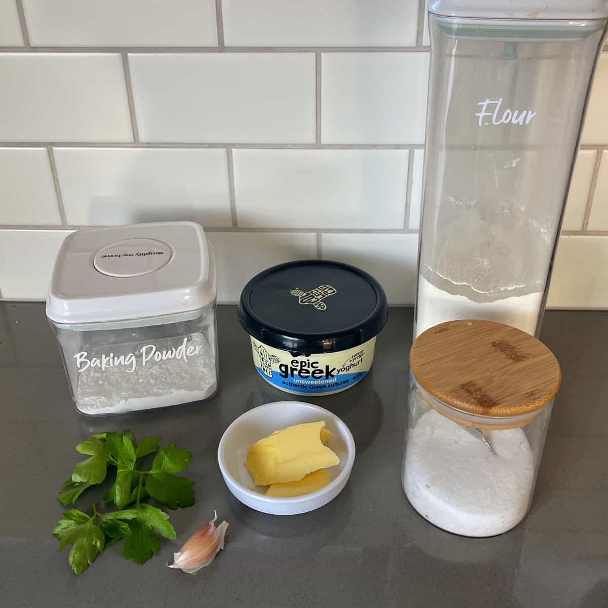 Four labeled containers of baking powder, flour, and sugar, along with Greek yogurt, parsley, a garlic clove, and a small dish of butter on a kitchen counter.
