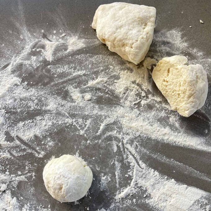 Three pieces of dough, one shaped into a ball, rest on a floured surface with scattered flour.
