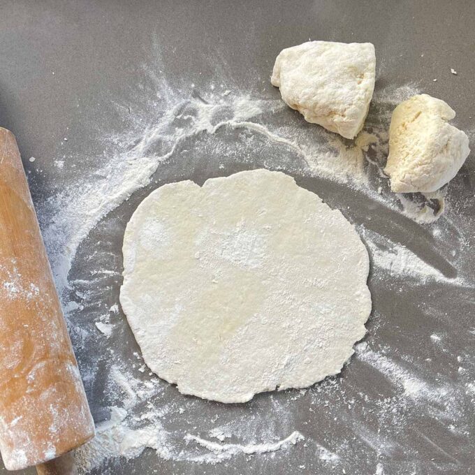 Round piece of dough rolled flat on a floured surface, with two unrolled dough pieces and a wooden rolling pin nearby.