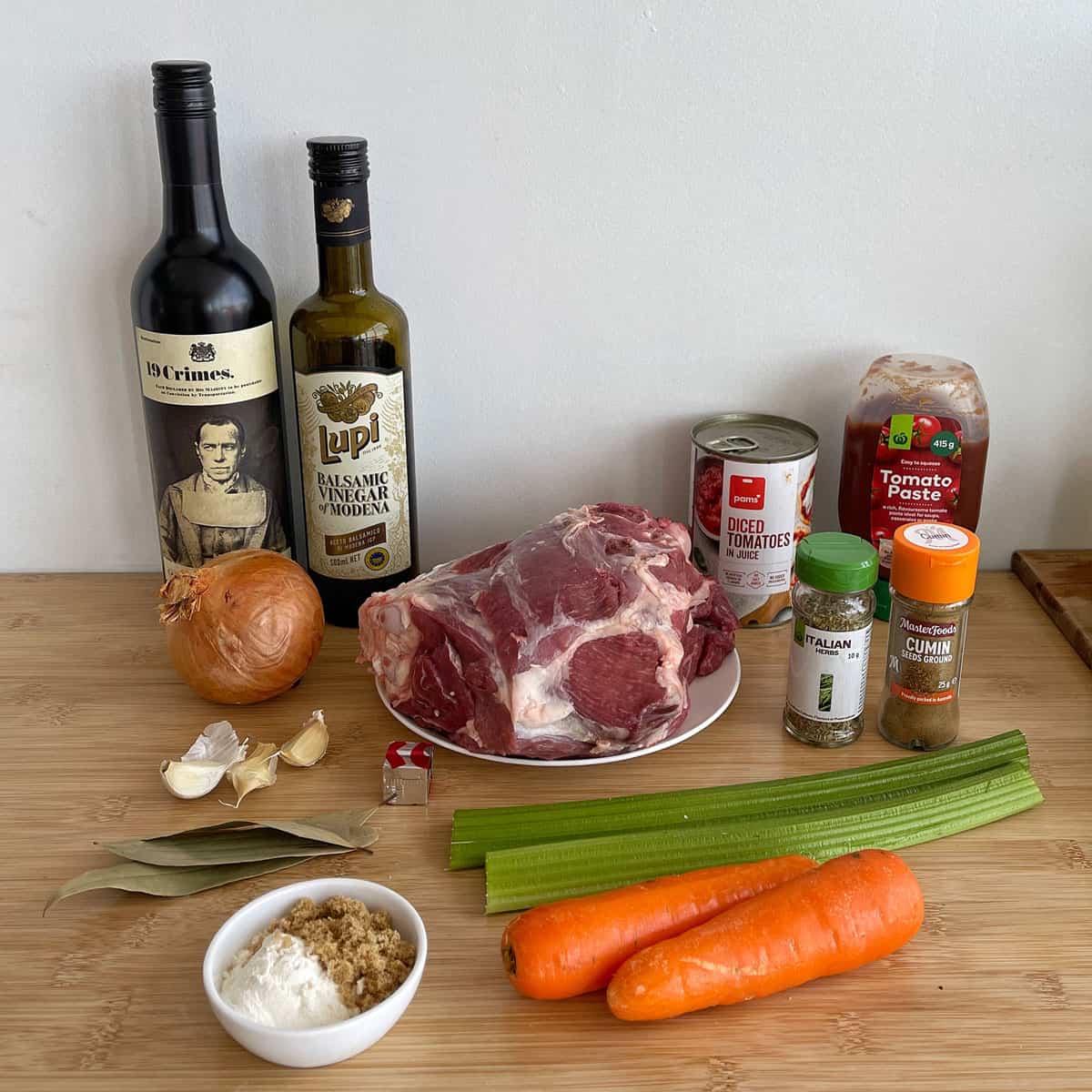 A selection of ingredients on a wooden surface, including a raw beef roast, wine, olive oil, vegetables, canned tomatoes, tomato paste, herbs, and spices.