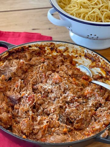 A pot of shredded beef in tomato sauce sits on a red cloth next to a colander filled with cooked spaghetti on a wooden table.