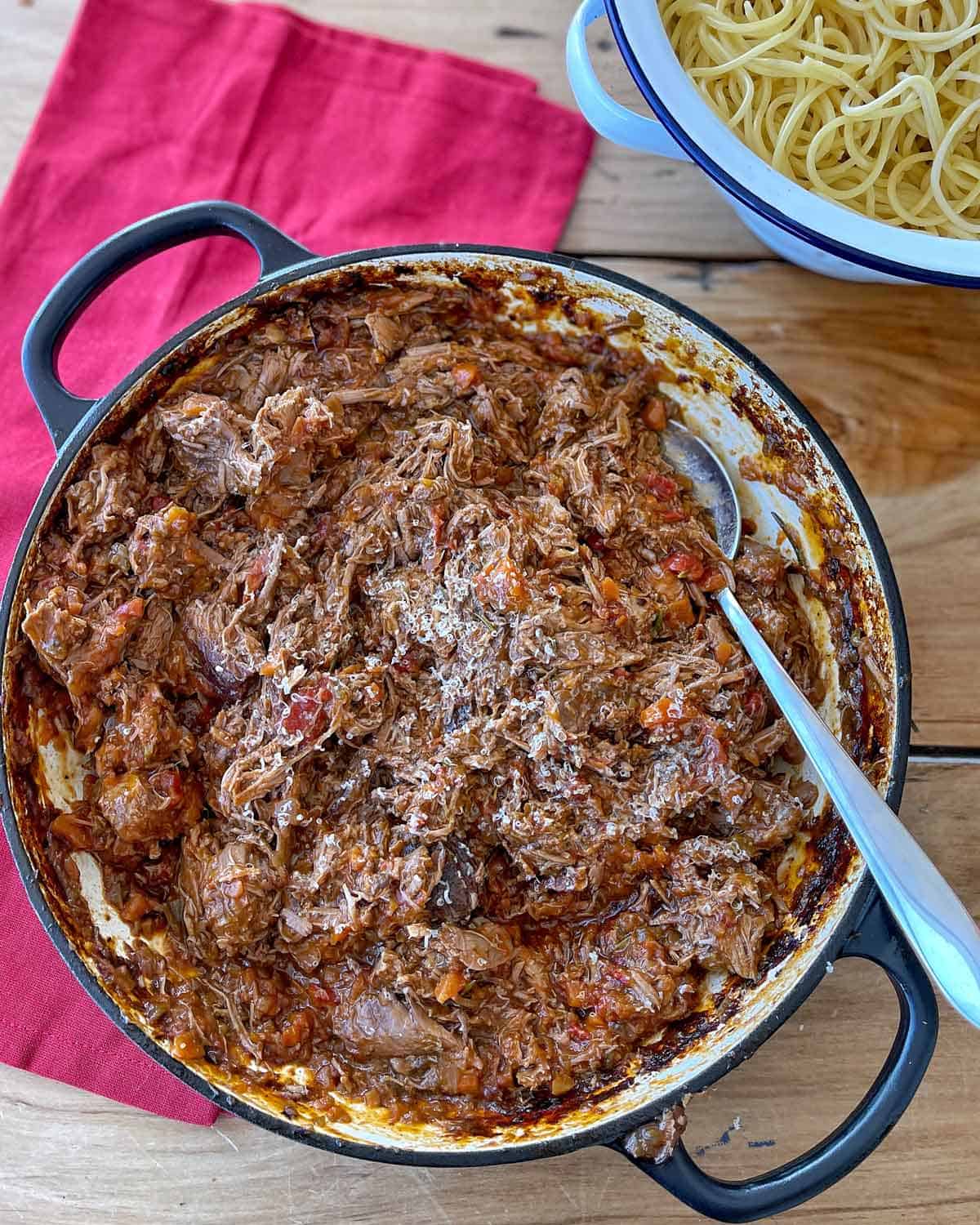 Lamb Ragu A pot of shredded beef ragu in sauce with a spoon, next to a bowl of cooked spaghetti on a wooden table with a red napkin.