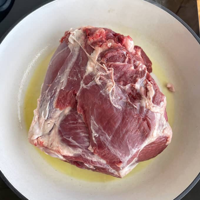 A large raw piece of red meat sits in a white pan with a thin layer of oil at the bottom.
