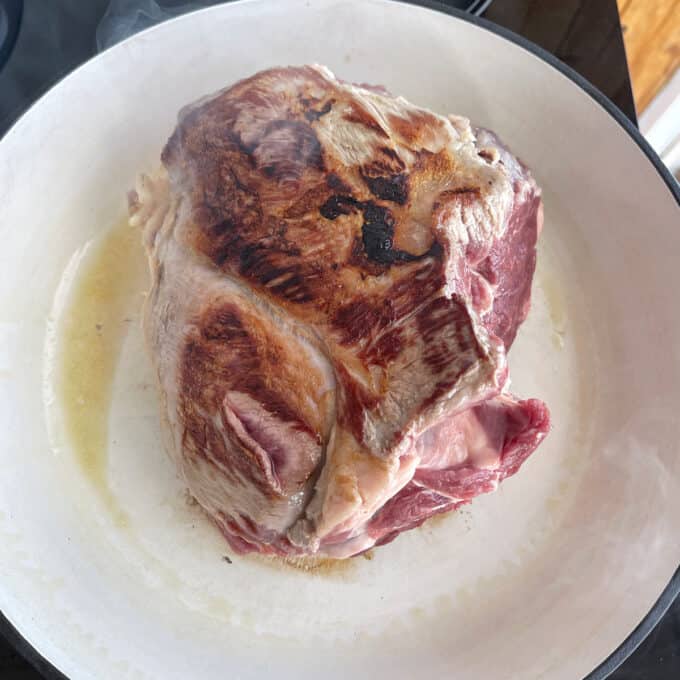 A large piece of meat is being seared in a white pan, showing browning on the surface and some raw areas remaining.