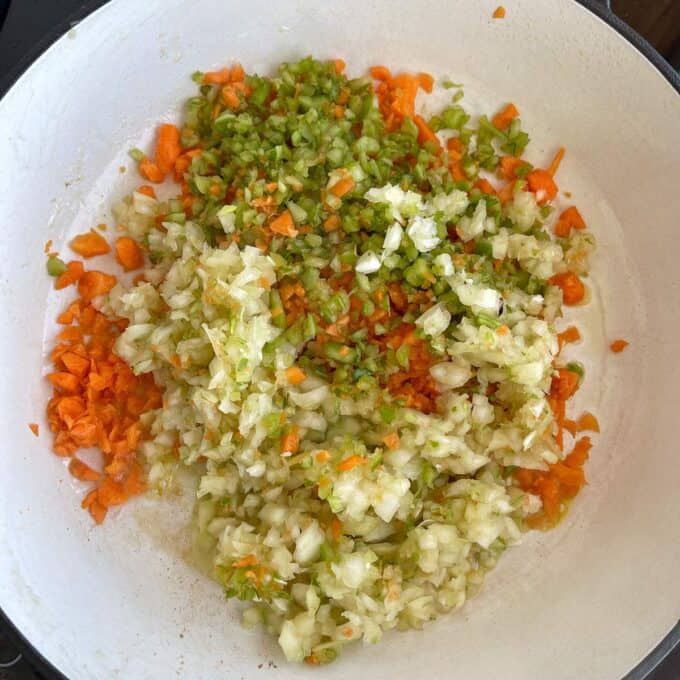 Chopped carrots, celery, and onions are sautéing in a white pan.