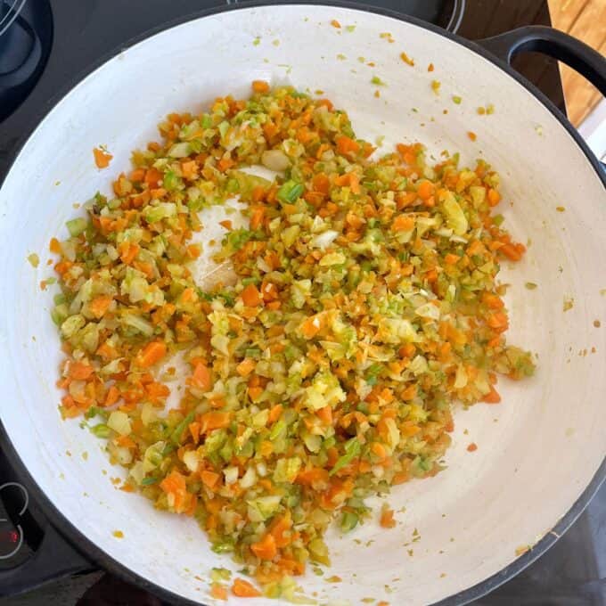 Chopped carrots, celery, and onions are sautéing in a large white pan on a stovetop.