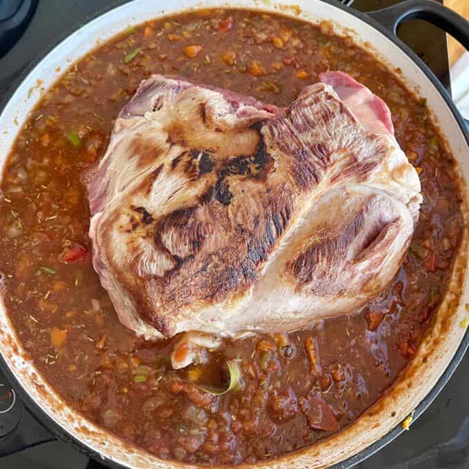A large browned roast sits on top of a pot of chunky tomato-based sauce with visible vegetables, cooking on a stovetop.