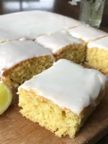A lemon sheet cake with a white icing glaze is cut into squares on a wooden board, with a lemon wedge placed beside the cake.