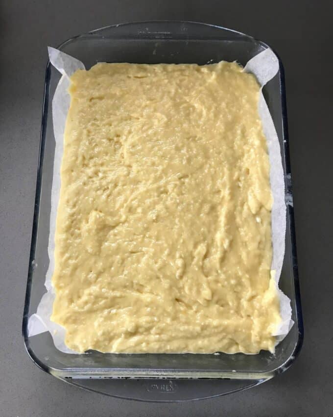 A glass baking dish lined with parchment paper, filled with an even layer of unbaked yellow batter on a dark countertop.