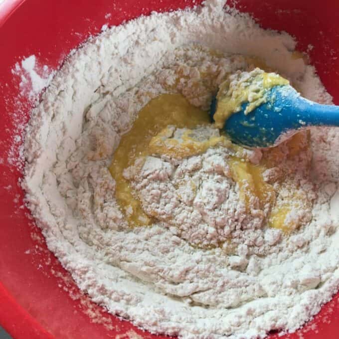 Flour and a wet mixture are being combined in a red bowl with a blue spatula, showing the beginning stages of mixing batter.