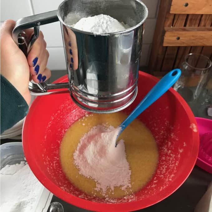 Hand holding a flour sifter over a red mixing bowl with batter; sifted flour is being added to the wet ingredients, and a blue spatula rests in the bowl.