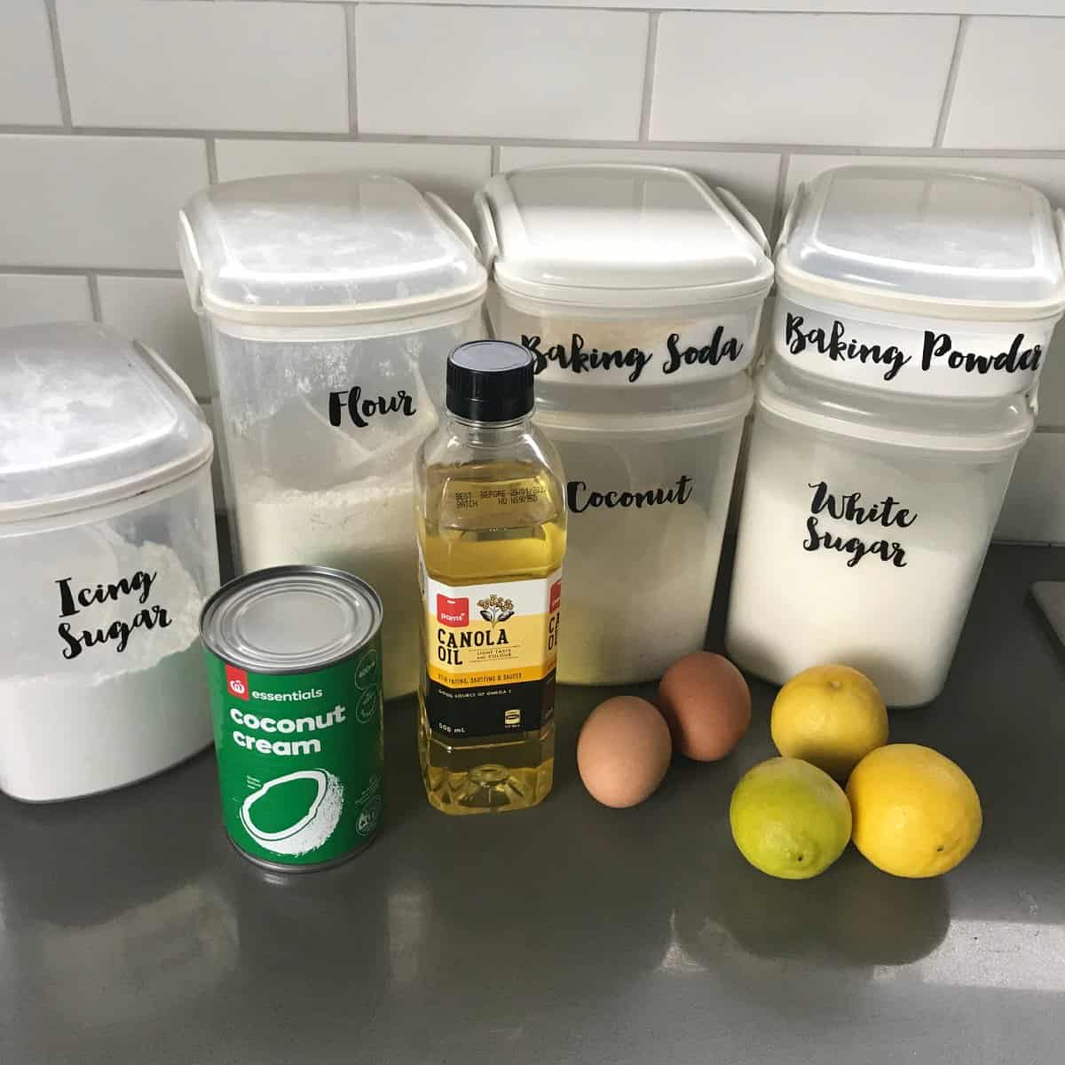 A countertop with containers labeled icing sugar, flour, baking soda, coconut, baking powder, and white sugar, plus a can of coconut cream, canola oil, three eggs, and two lemons.