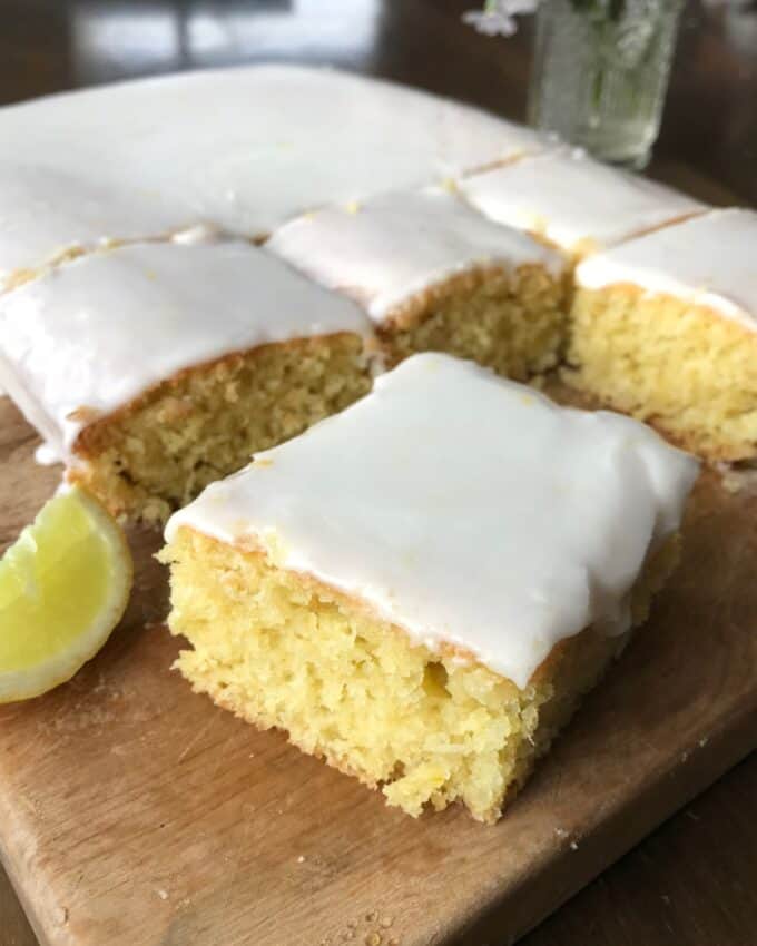 Lemon sheet cake with white icing cut into squares, displayed on a wooden board with a lemon wedge nearby.
