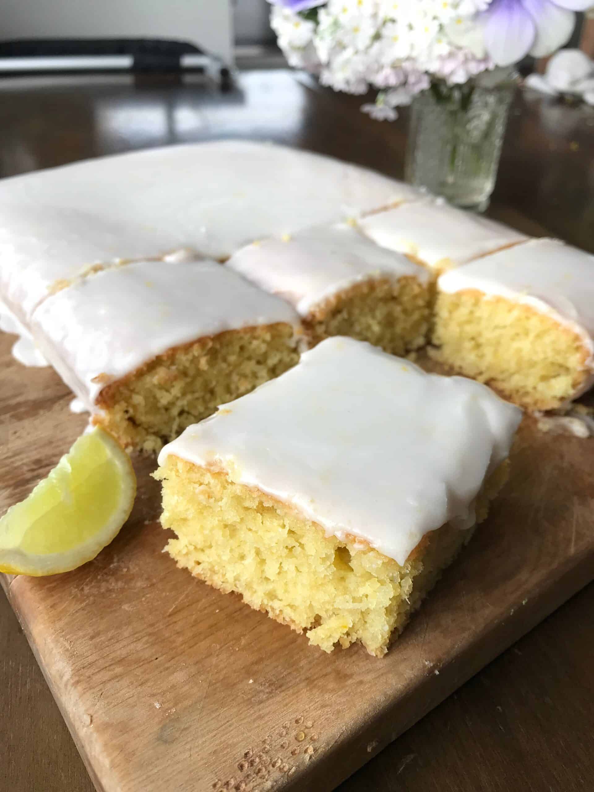 Lime and Coconut Cake A rectangular lemon cake with white icing is cut into squares on a wooden board, with a lemon wedge and a vase of flowers in the background.