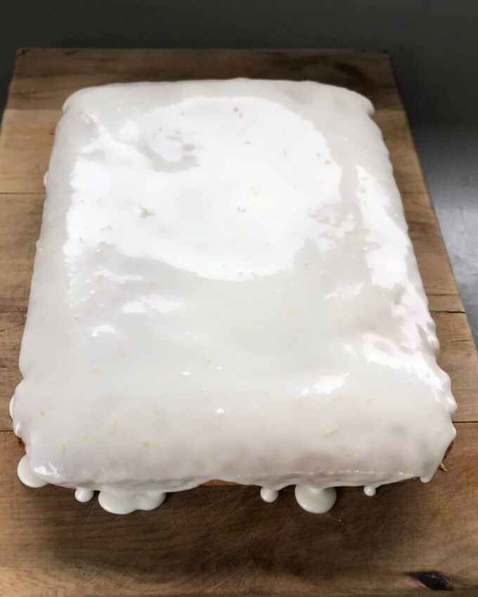 A rectangular cake on a wooden surface, covered with a smooth white glaze that drips slightly over the edges.