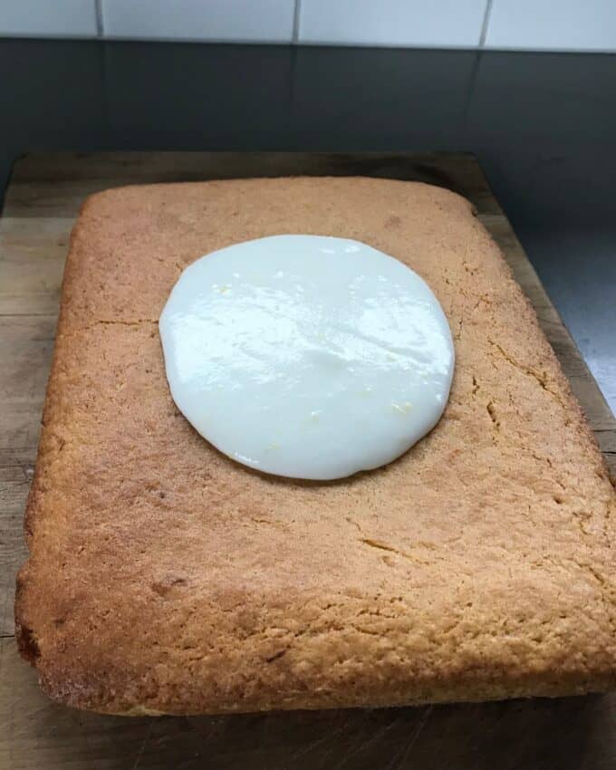A rectangular sponge cake on a wooden board with a round pool of white icing in the center.