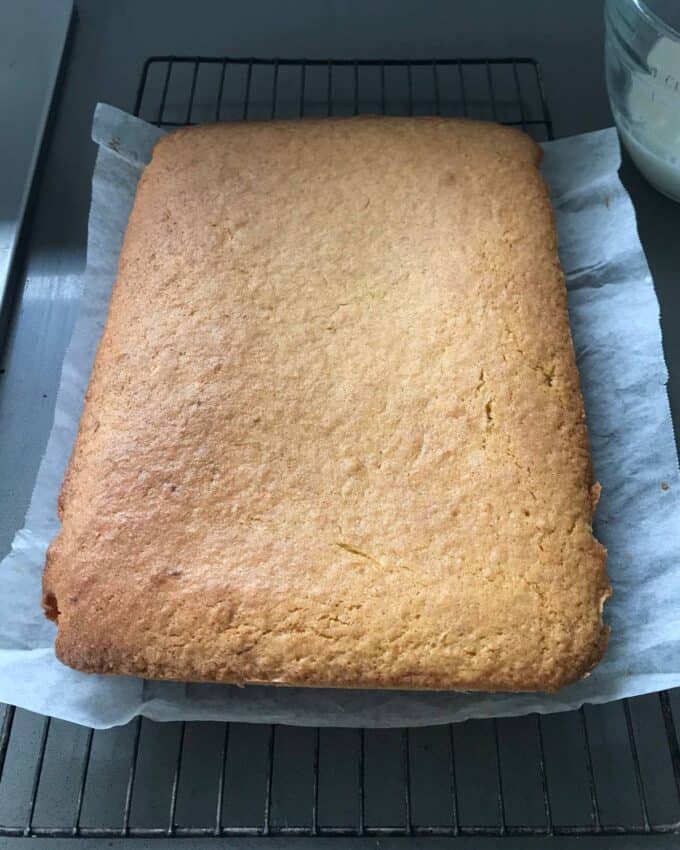 A rectangular sheet cake with a golden-brown top cools on a wire rack, lined with parchment paper.