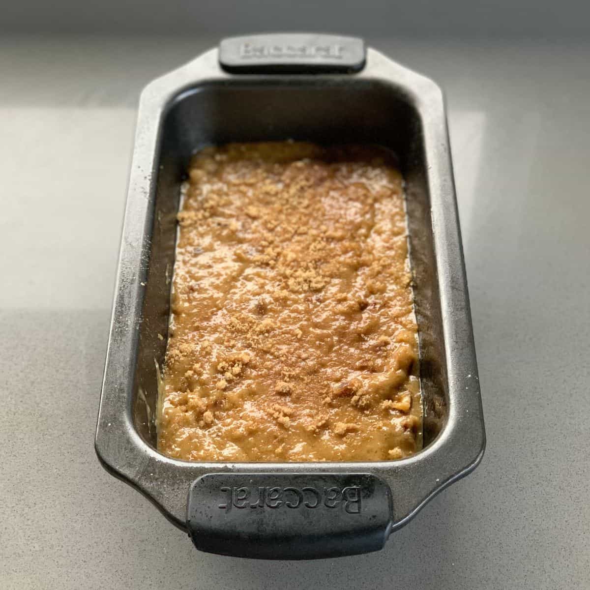 A metal loaf pan filled with unbaked batter, sprinkled with crumb topping, sits on a light-colored countertop.