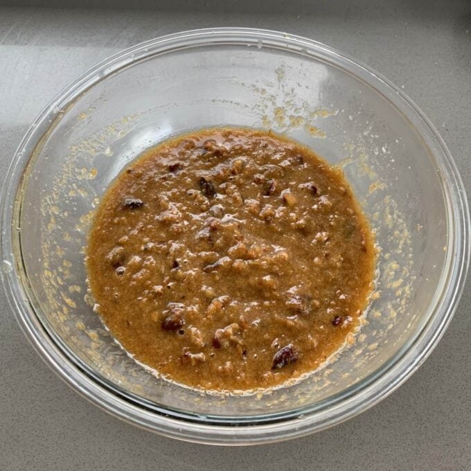 A glass bowl containing a thick brown batter mixture with visible chunks, placed on a grey countertop.