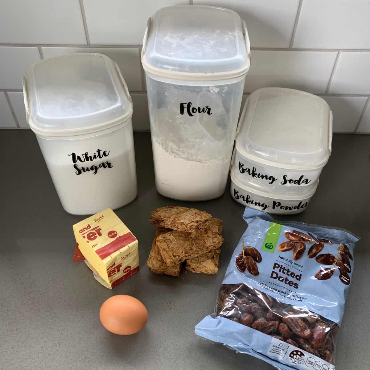 Ingredients on a kitchen counter: containers of white sugar, flour, and baking soda/powder, a block of butter, one egg, Weet-Bix biscuits, and a bag of pitted dates.