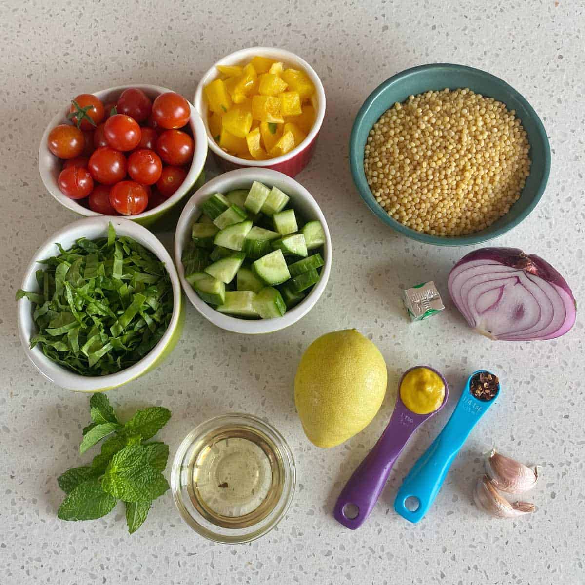 Assorted ingredients on a countertop: cherry tomatoes, yellow bell pepper, couscous, cucumber, greens, onion, mint, lemon, oil, mustard, chili flakes, garlic, and a bouillon cube.