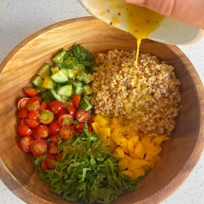 A wooden bowl filled with chopped cucumber, cherry tomatoes, yellow bell pepper, spinach, and cooked couscous, with dressing being poured over the top.