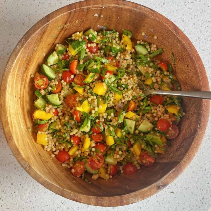A wooden bowl filled with couscous salad containing cherry tomatoes, cucumber, yellow bell pepper, and fresh herbs, with a spoon on the side.