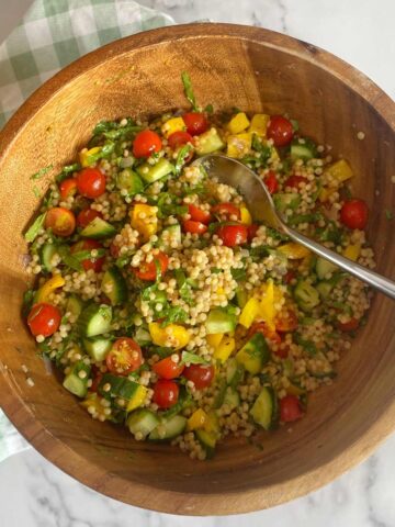A wooden bowl filled with couscous salad, cherry tomatoes, cucumber, yellow bell pepper, and herbs, with a serving spoon inside. A green-and-white cloth is underneath the bowl.