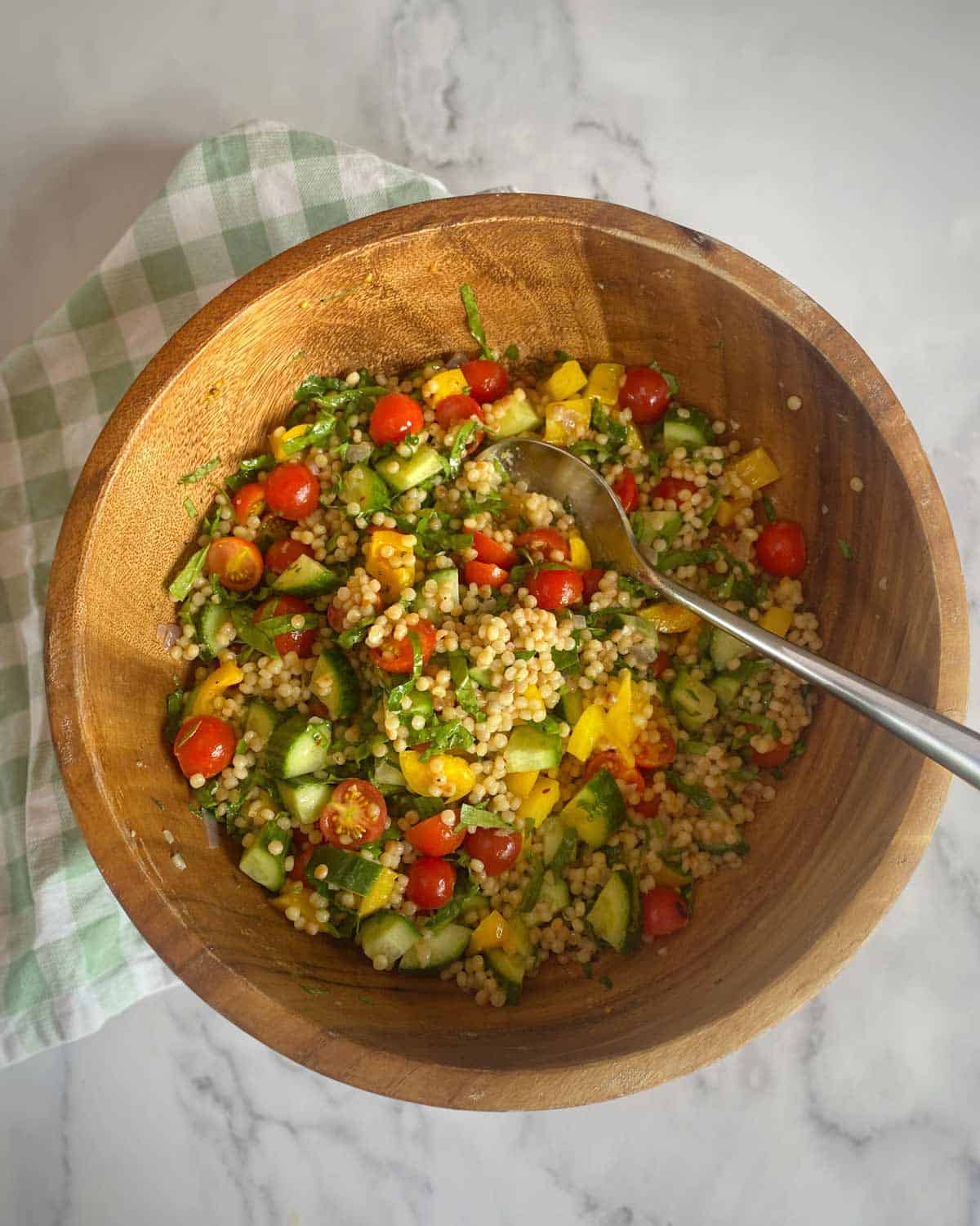 Pearl Couscous Salad A wooden bowl filled with couscous salad, including cherry tomatoes, cucumbers, yellow bell peppers, and herbs, with a spoon inside. The bowl rests on a green checkered cloth.