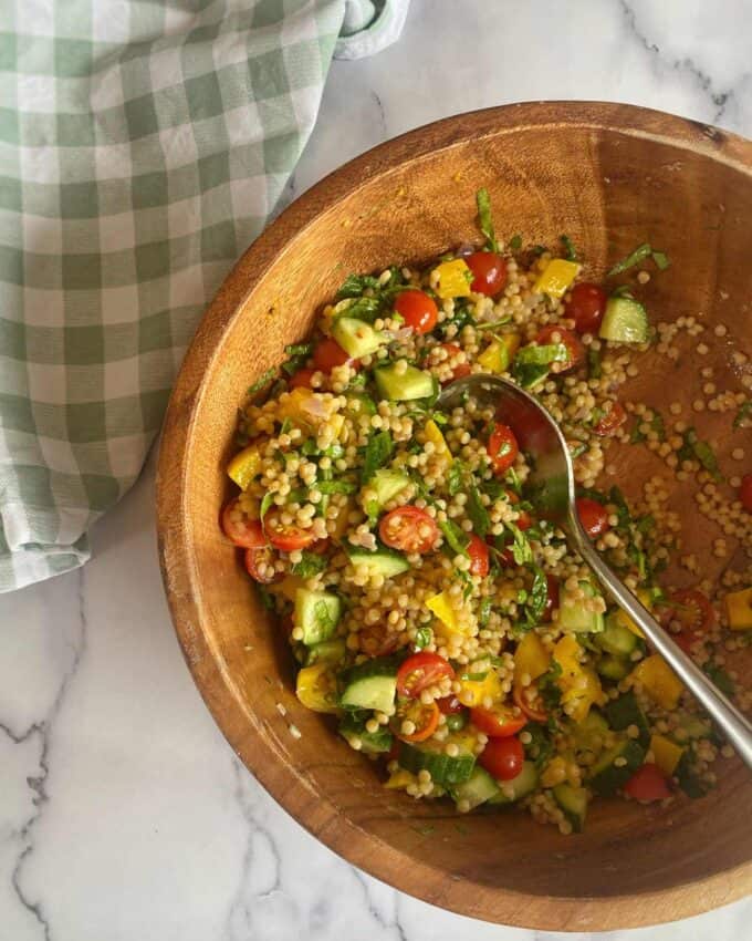 A wooden bowl filled with couscous salad containing cherry tomatoes, cucumber, yellow pepper, and herbs, with a metal serving spoon. A green checkered cloth is nearby.