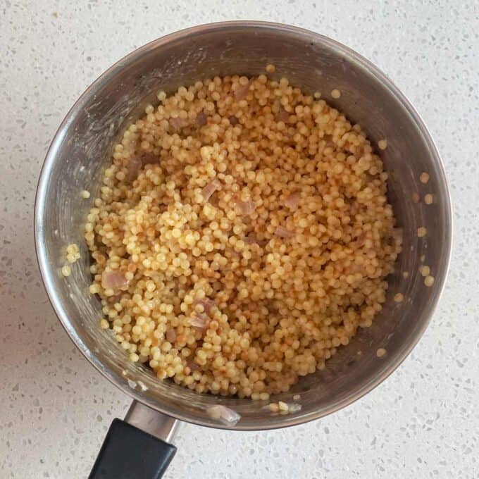 A pot of cooked Israeli couscous mixed with small pieces of onion sits on a white countertop.