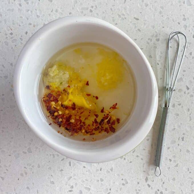 A white bowl containing oil, minced garlic, grated ginger, red pepper flakes, and a yellow paste, with a small metal whisk beside it on a speckled countertop.