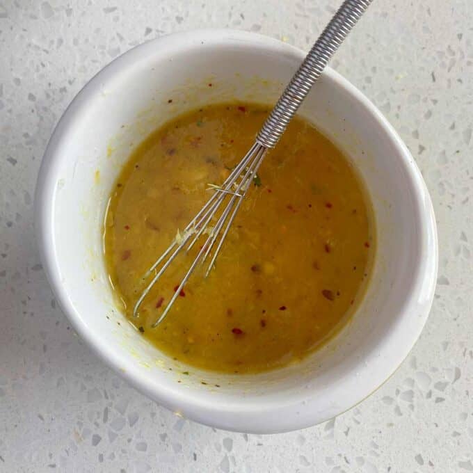 A small whisk sits in a white bowl containing a yellow, slightly chunky liquid with visible spices on a speckled countertop.