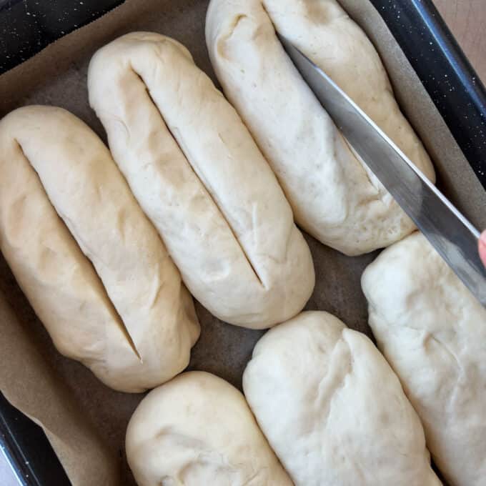 A hand slices slits into unbaked bread dough loaves arranged on a parchment-lined baking tray.