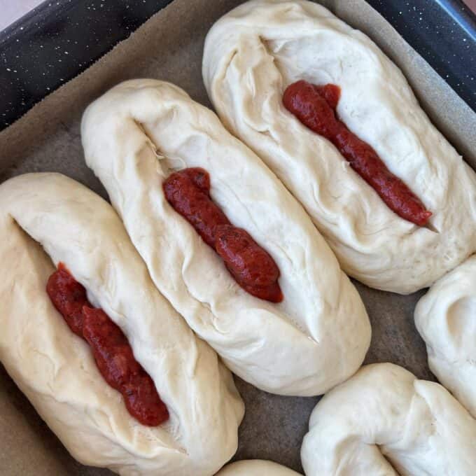 Three unbaked bread rolls on a parchment-lined tray, each with a line of red tomato sauce in the center.