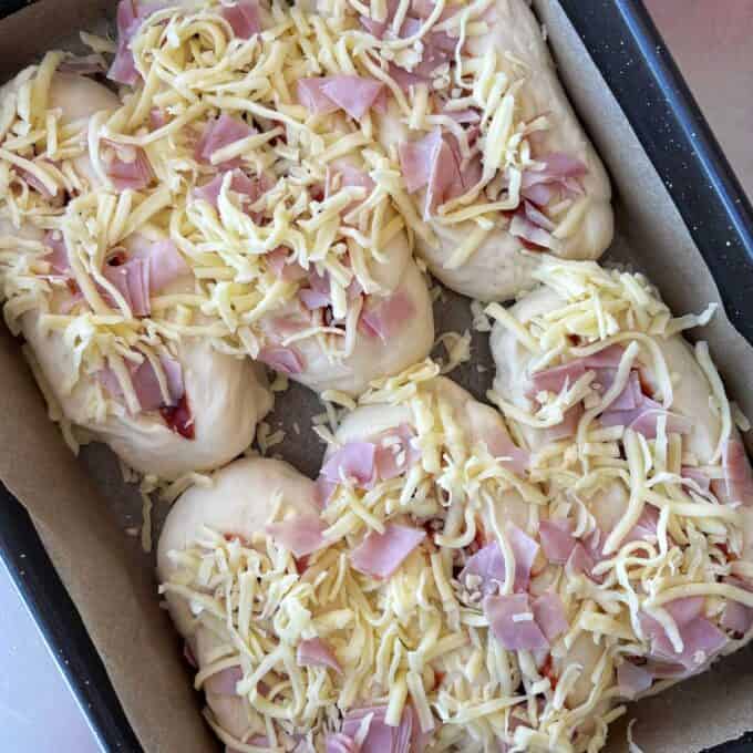 A baking tray lined with parchment paper holds unbaked bread rolls topped with shredded cheese and chopped ham.