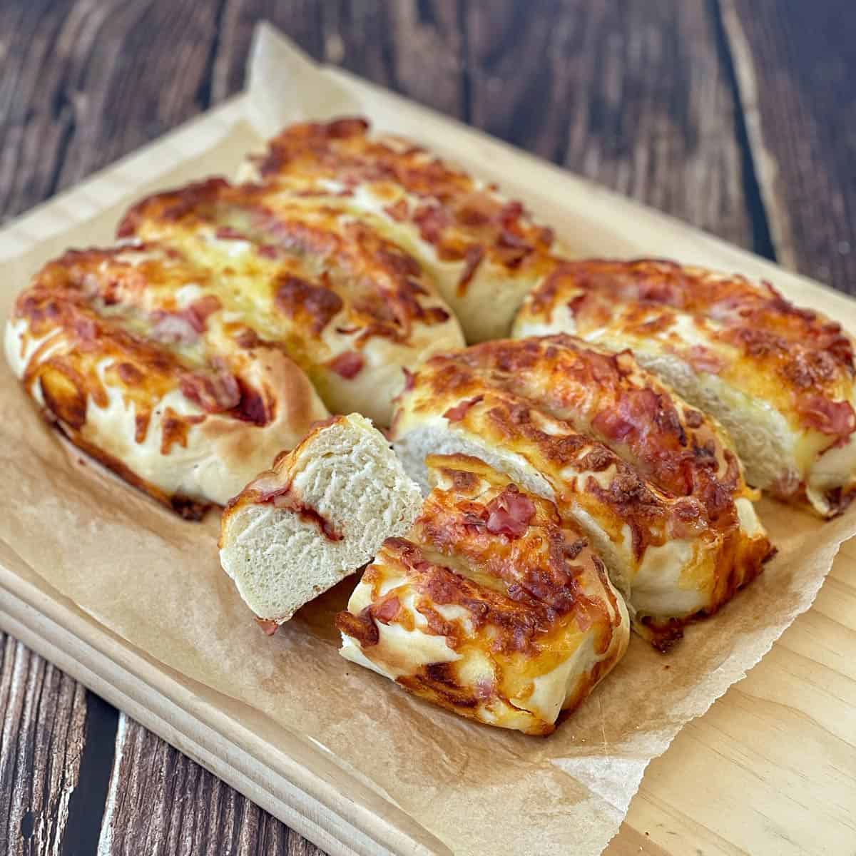 A sliced, baked bread loaf topped with melted cheese and bits of ham sits on parchment paper atop a wooden board.