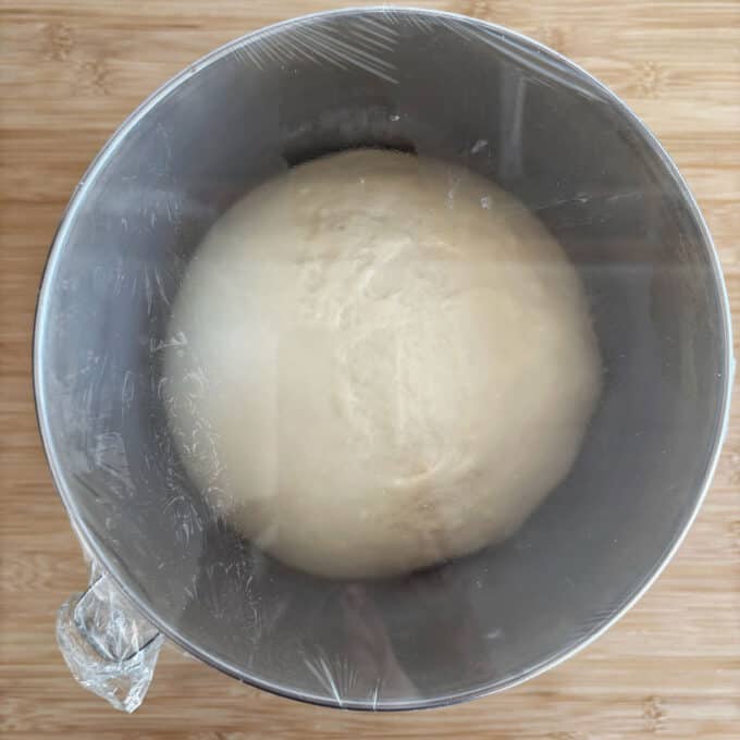 A ball of dough rises in a metal mixing bowl covered with plastic wrap, placed on a wooden surface.