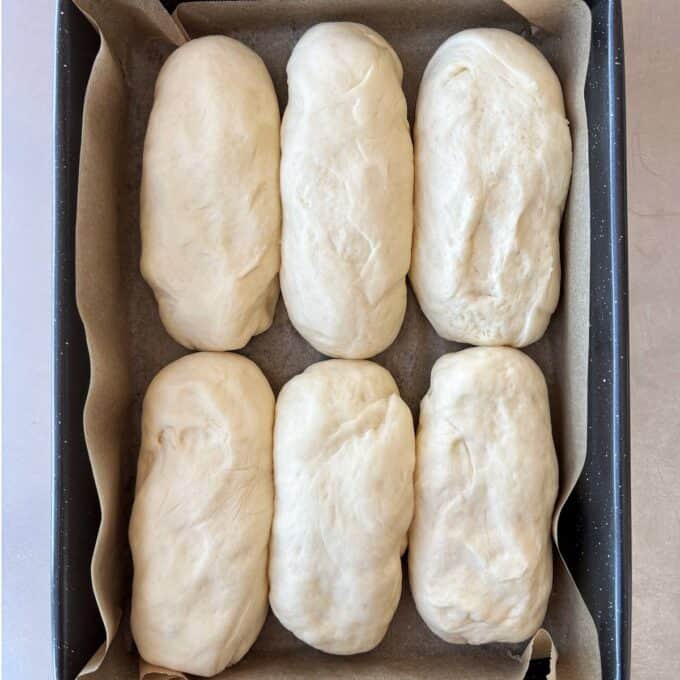 Six unbaked bread rolls arranged in two rows on parchment paper in a rectangular baking tray.