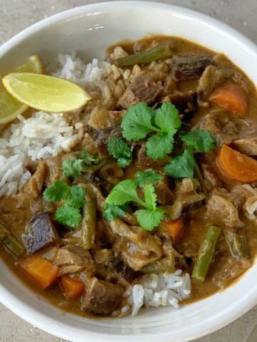 A bowl of white rice topped with beef curry containing carrots, green beans, and eggplant, garnished with fresh cilantro and two lemon wedges.