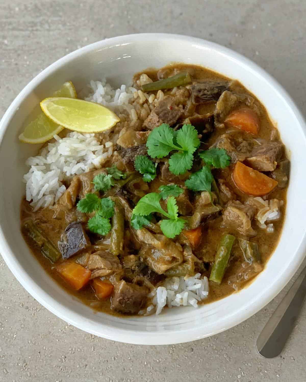 beef red curry A bowl of rice topped with beef curry, vegetables like carrots and green beans, garnished with cilantro and lemon wedges.